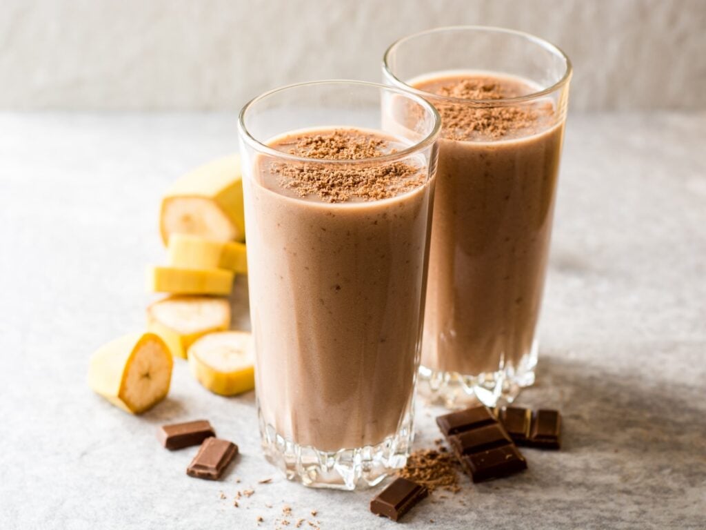 Two glasses of chocolate banana smoothie topped with grated chocolate, with banana slices and chocolate pieces on a light surface.