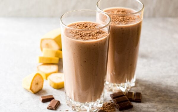 Two glasses of chocolate banana smoothie topped with grated chocolate, with banana slices and chocolate pieces on a light surface.