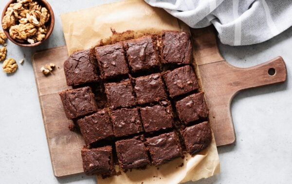 A batch of chocolate brownies cut into squares sits on baking parchment atop a wooden board, with a small bowl of walnuts nearby and a striped cloth in the corner.