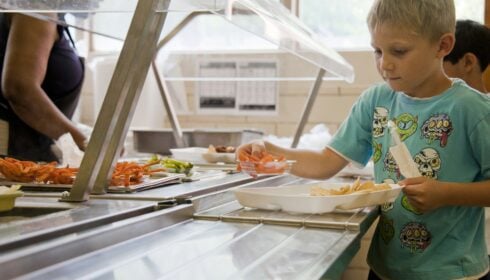 public food. A young boy in a blue shirt picks food from a canteen serving line, placing items onto a tray. Another person serves food in the background.