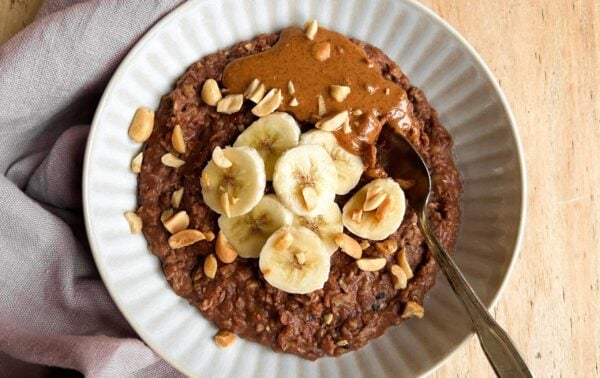 Bowl of chocolate porridge topped with banana slices, peanut butter, and chopped peanuts, with a spoon resting inside.