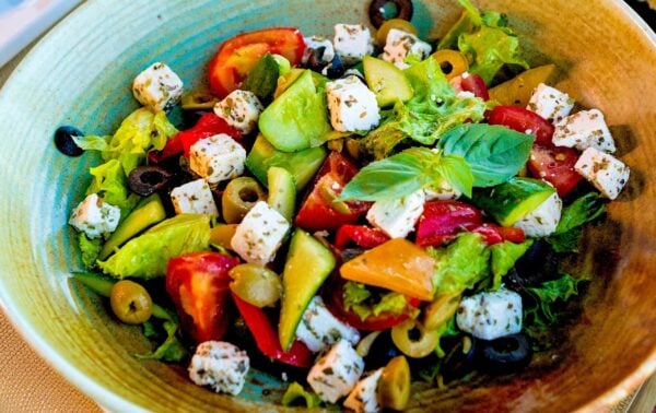 A bowl of Greek salad with lettuce, tomatoes, cucumbers, olives, and feta cheese cubes, garnished with fresh basil.