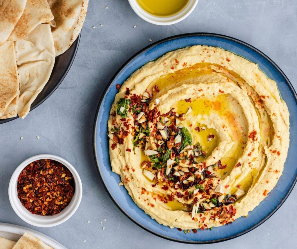 A blue plate of houmous topped with herbs, chopped nuts, and olive oil, surrounded by pitta bread, chilli flakes, and a small bowl of olive oil.