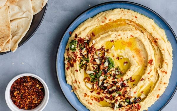 A blue plate of houmous topped with herbs, chopped nuts, and olive oil, surrounded by pitta bread, chilli flakes, and a small bowl of olive oil.
