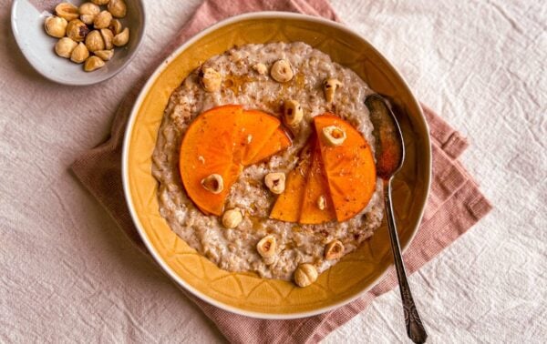 A bowl of porridge topped with persimmon slices and hazelnuts, with a spoon in the bowl and a small dish of hazelnuts beside it on a textured serviette.