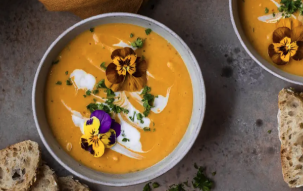 Bowl of creamy orange soup garnished with edible flowers, herbs, and a swirl of cream, served with slices of wholegrain bread on the side.