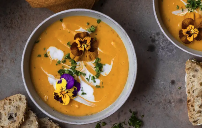 Bowl of creamy orange soup garnished with edible flowers, herbs, and a swirl of cream, served with slices of wholegrain bread on the side.
