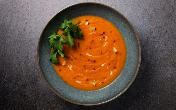 A bowl of orange soup garnished with fresh coriander, cream swirls, and red chilli flakes, placed on a grey stone surface.