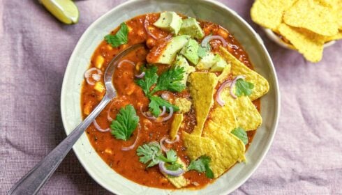 A bowl of chilli topped with avocado, red onion, coriander, and tortilla chips, with lime wedges and extra crisps on the side.