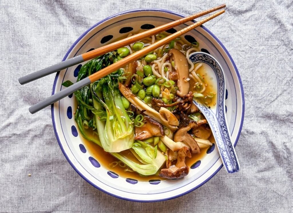 Bowl of vegetable ramen with pak choi, mushrooms, edamame, and spring onions in broth, served with chopsticks and a patterned spoon on a light cloth surface.