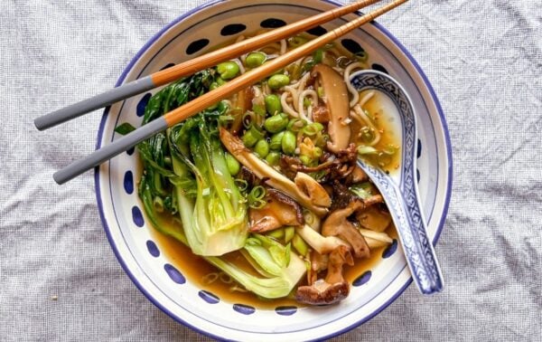 Bowl of vegetable ramen with pak choi, mushrooms, edamame, and spring onions in broth, served with chopsticks and a patterned spoon on a light cloth surface.