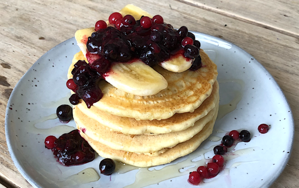 A stack of pancakes topped with banana slices, berry compote, and syrup, served on a light grey plate on a wooden table.