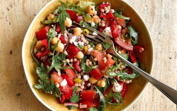 A bowl of salad with chickpeas, couscous, rocket, red peppers, tomatoes, feta cheese, and onions, with a fork and spoon on a wooden surface.