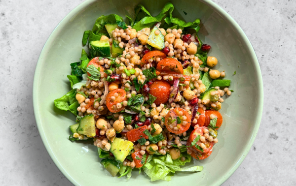 A bowl of salad with couscous, chickpeas, cherry tomatoes, avocado, chopped greens, and herbs on a light grey surface.