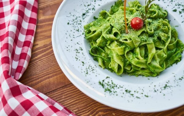 A plate of green fettuccine pasta garnished with grated cheese, a cherry tomato, herbs, and a sprig of rosemary, with a red and white checked serviette beside it on a wooden table.