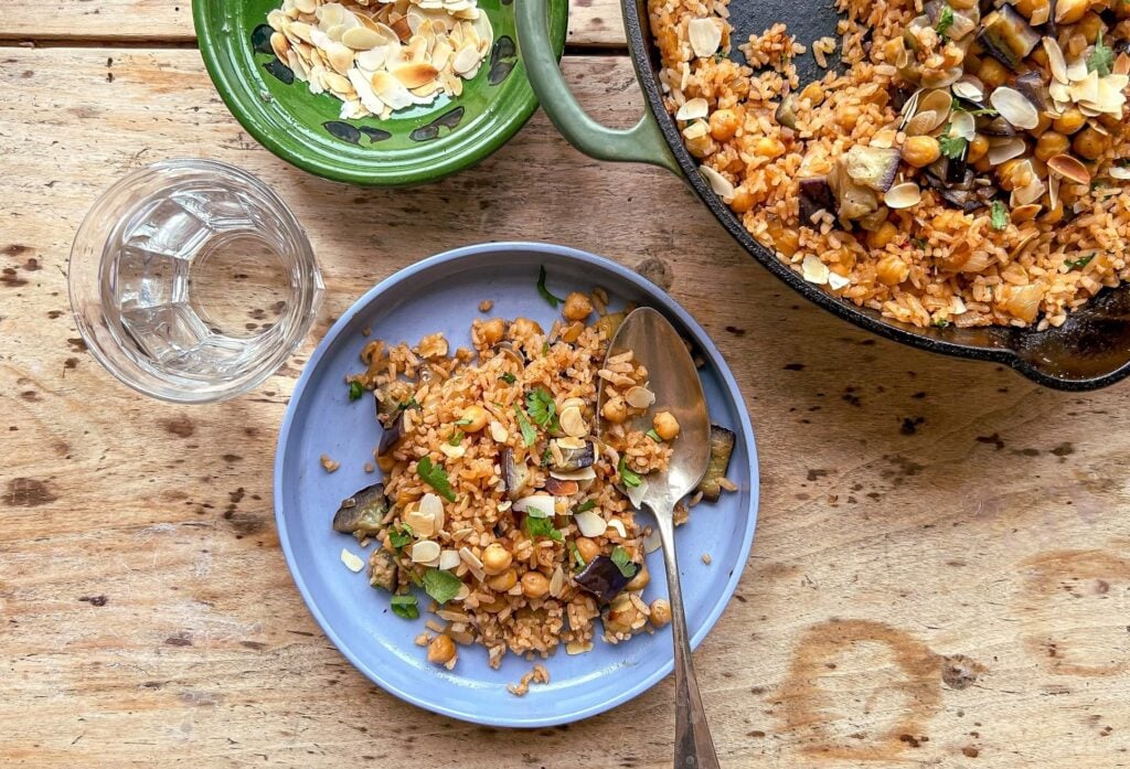 A blue plate with rice, chickpeas, aubergine, and herbs beside a spoon, a glass of water, a bowl of almond slices, and a pan with more rice mixture on a wooden table.