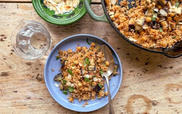 A blue plate with rice, chickpeas, aubergine, and herbs beside a spoon, a glass of water, a bowl of almond slices, and a pan with more rice mixture on a wooden table.