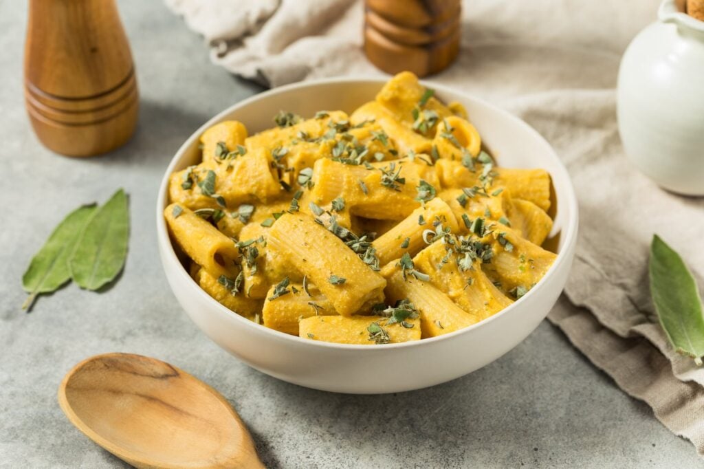 A white bowl filled with creamy rigatoni pasta topped with fresh herbs, placed on a light surface with a wooden spoon, bay leaves, and wooden shakers nearby.