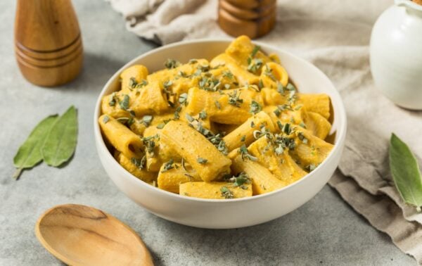 A white bowl filled with creamy rigatoni pasta topped with fresh herbs, placed on a light surface with a wooden spoon, bay leaves, and wooden shakers nearby.