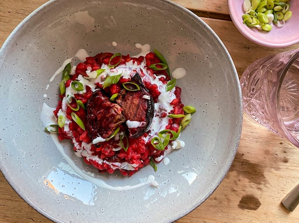 A bowl of beetroot risotto topped with roasted beetroot, sliced spring onions, and a drizzle of white sauce, placed on a wooden table next to a pink dish of spring onions and a glass of water.