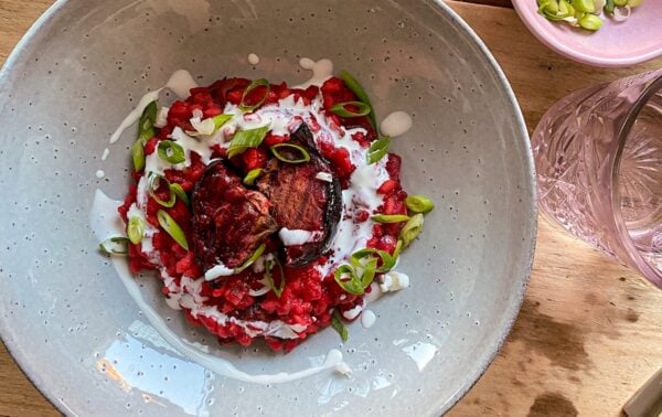 A bowl of beetroot risotto topped with roasted beetroot, sliced spring onions, and a drizzle of white sauce, placed on a wooden table next to a pink dish of spring onions and a glass of water.