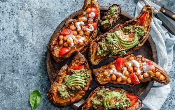 A wooden plate with roasted aubergine halves topped with chickpeas, avocado, tomatoes, greens, and drizzled sauce, on a grey surface with cutlery and a serviette nearby.