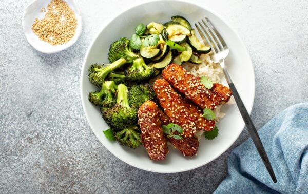 A white bowl with rice, glazed tofu topped with sesame seeds, roasted broccoli, courgette, and fresh herbs, with a fork resting on the rim. A small dish of sesame seeds is nearby.