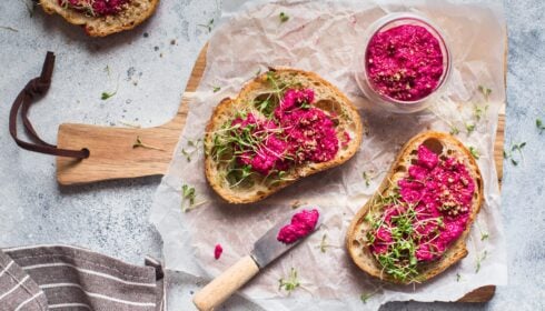 Two slices of bread with beetroot spread and sprouts on baking parchment, with a small bowl of beetroot spread and a knife on a wooden board.