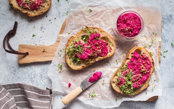 Two slices of bread with beetroot spread and sprouts on baking parchment, with a small bowl of beetroot spread and a knife on a wooden board.