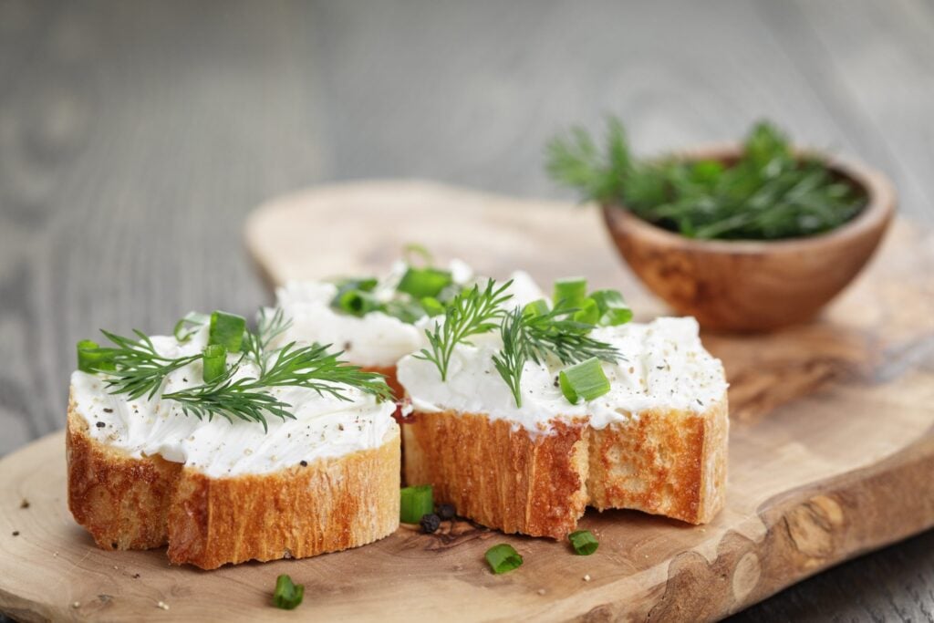 Two slices of bread topped with cream cheese, chopped spring onions, and dill are on a wooden board. A small bowl with extra herbs is in the background.