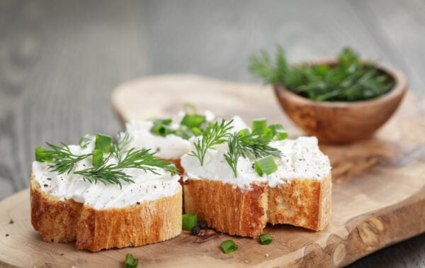 Two slices of bread topped with cream cheese, chopped spring onions, and dill are on a wooden board. A small bowl with extra herbs is in the background.