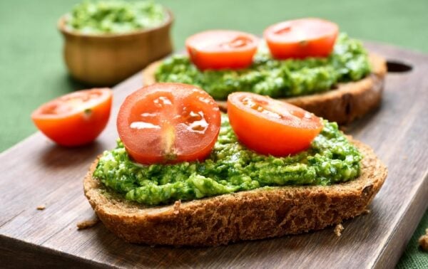 Two slices of wholegrain bread topped with green pesto and halved cherry tomatoes on a wooden board, with a small bowl of pesto in the background.