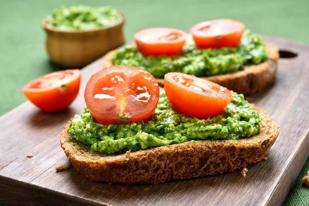 Two slices of wholegrain bread topped with green pesto and halved cherry tomatoes on a wooden board, with a small bowl of pesto in the background.