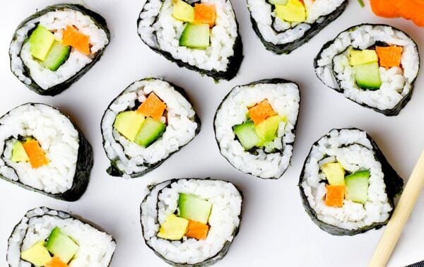 Nine vegetable sushi rolls with rice, cucumber, carrot, and avocado are arranged on a white surface next to chopsticks.