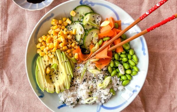 A bowl with rice, sliced avocado, sweetcorn, cucumber, carrots, edamame, spring onions, and black sesame seeds, with chopsticks and a dish of soy sauce on the side.