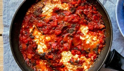 A black frying pan filled with stewed tomatoes, greens, and poached eggs sits on a blue textured cloth, with part of a blue plate visible at the side.