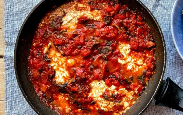 A black frying pan filled with stewed tomatoes, greens, and poached eggs sits on a blue textured cloth, with part of a blue plate visible at the side.