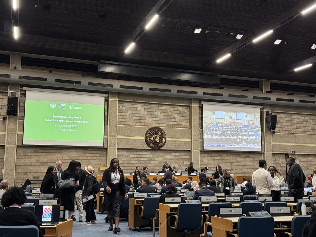 SBI-7. Delegates gather and interact in a large conference room with two screens displaying event information and a meeting photo at the front.