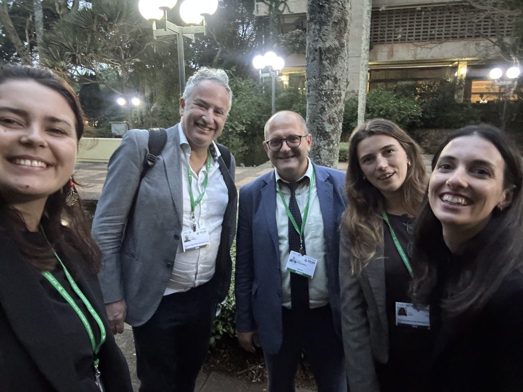 Five people wearing conference badges and business attire stand outdoors, smiling at the camera in a garden setting with trees and lights visible in the background.