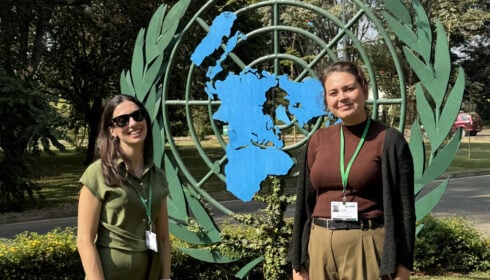 SBI-7. Two women with green lanyards stand in front of a United Nations emblem outdoors, surrounded by greenery and trees.
