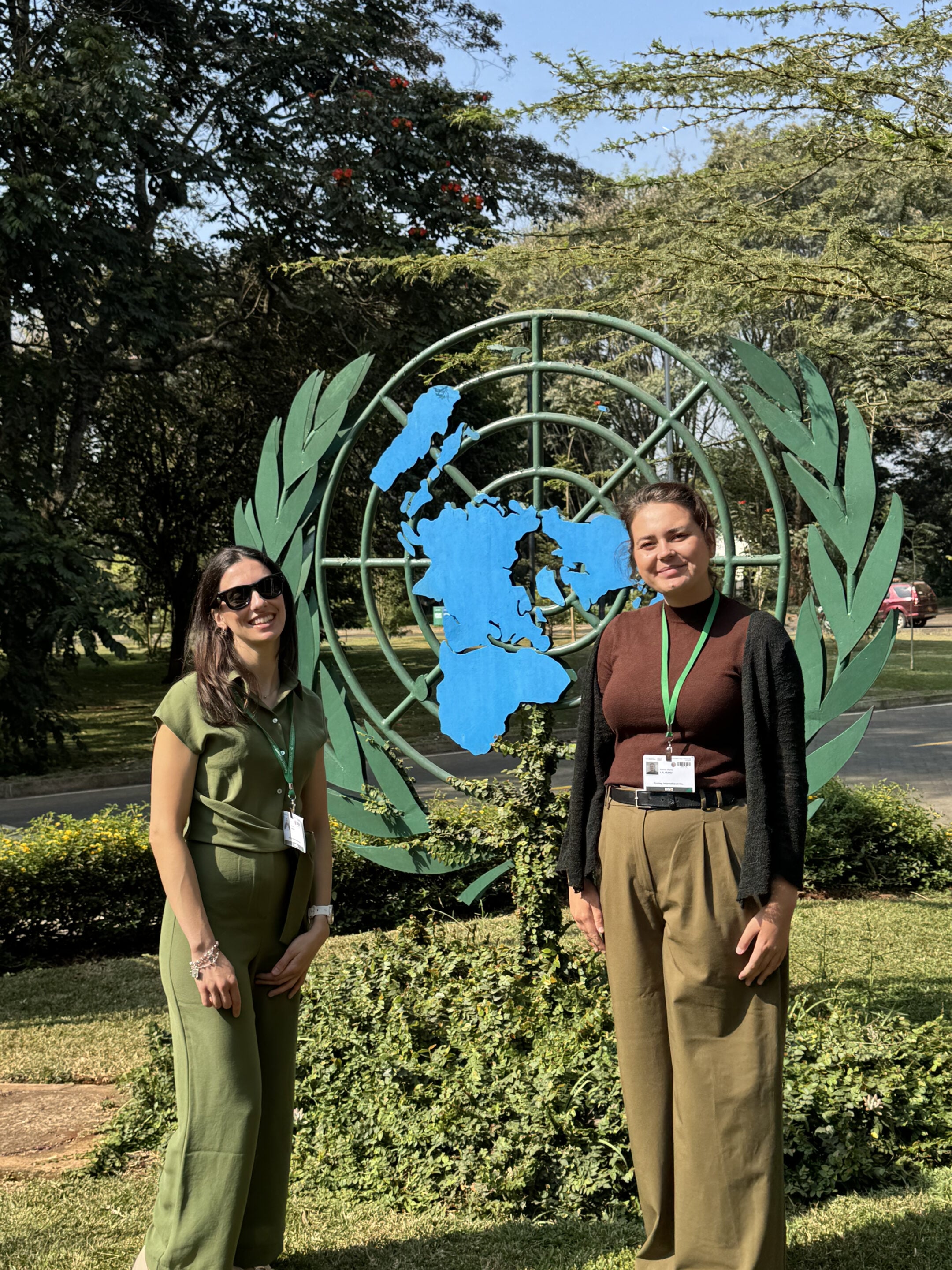 SBI-7. Two women with green lanyards stand in front of a United Nations emblem outdoors, surrounded by greenery and trees.