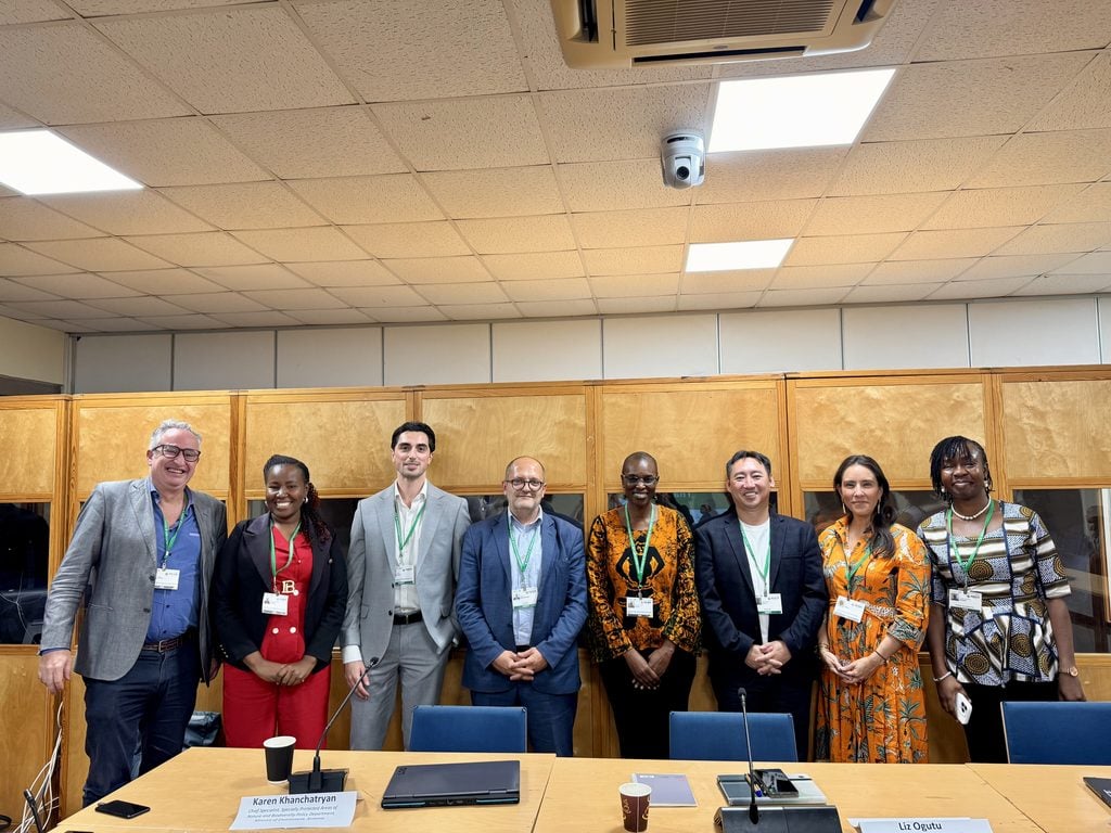 SBI-7. Eight people standing and posing for a group photo in a conference room, wearing business or business-casual attire and conference badges.