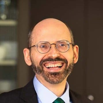 A man with glasses, a beard, and a bald head is smiling whilst wearing a suit and tie.