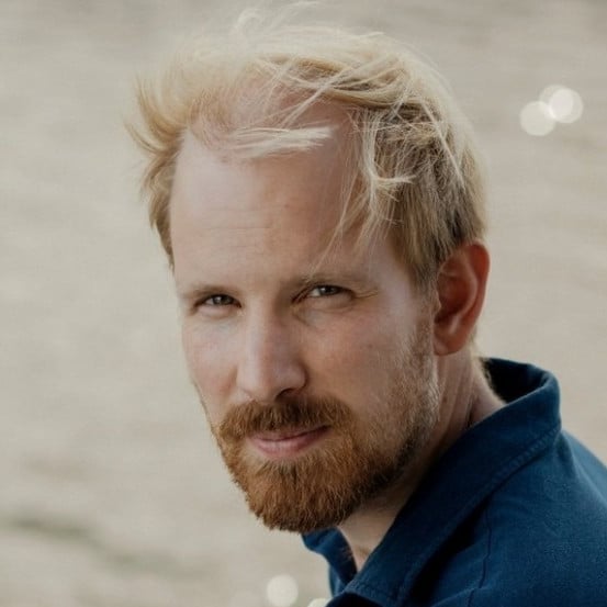 A man with light skin, blond hair, and a reddish beard looks at the camera. He is outdoors, wearing a blue shirt, with a blurred, neutral background.