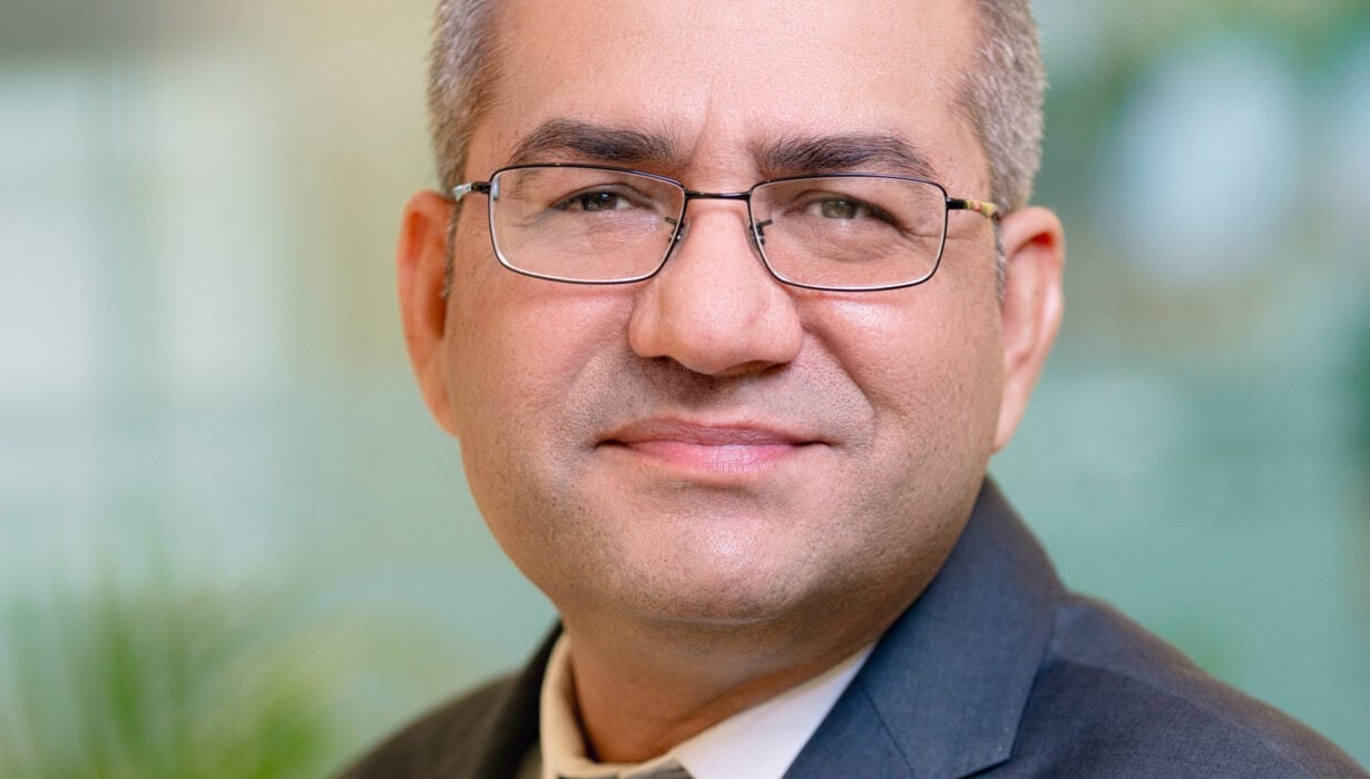 A middle-aged man with short grey hair, glasses, and a suit smiles slightly at the camera in an indoor setting with a blurred background.