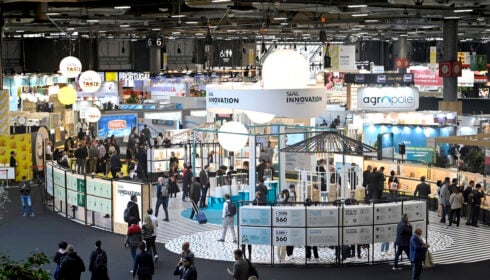 Large indoor trade fair with multiple stands, informational displays, and people walking and interacting in a well-lit exhibition hall. Signs with company names and bright overhead lights are visible.