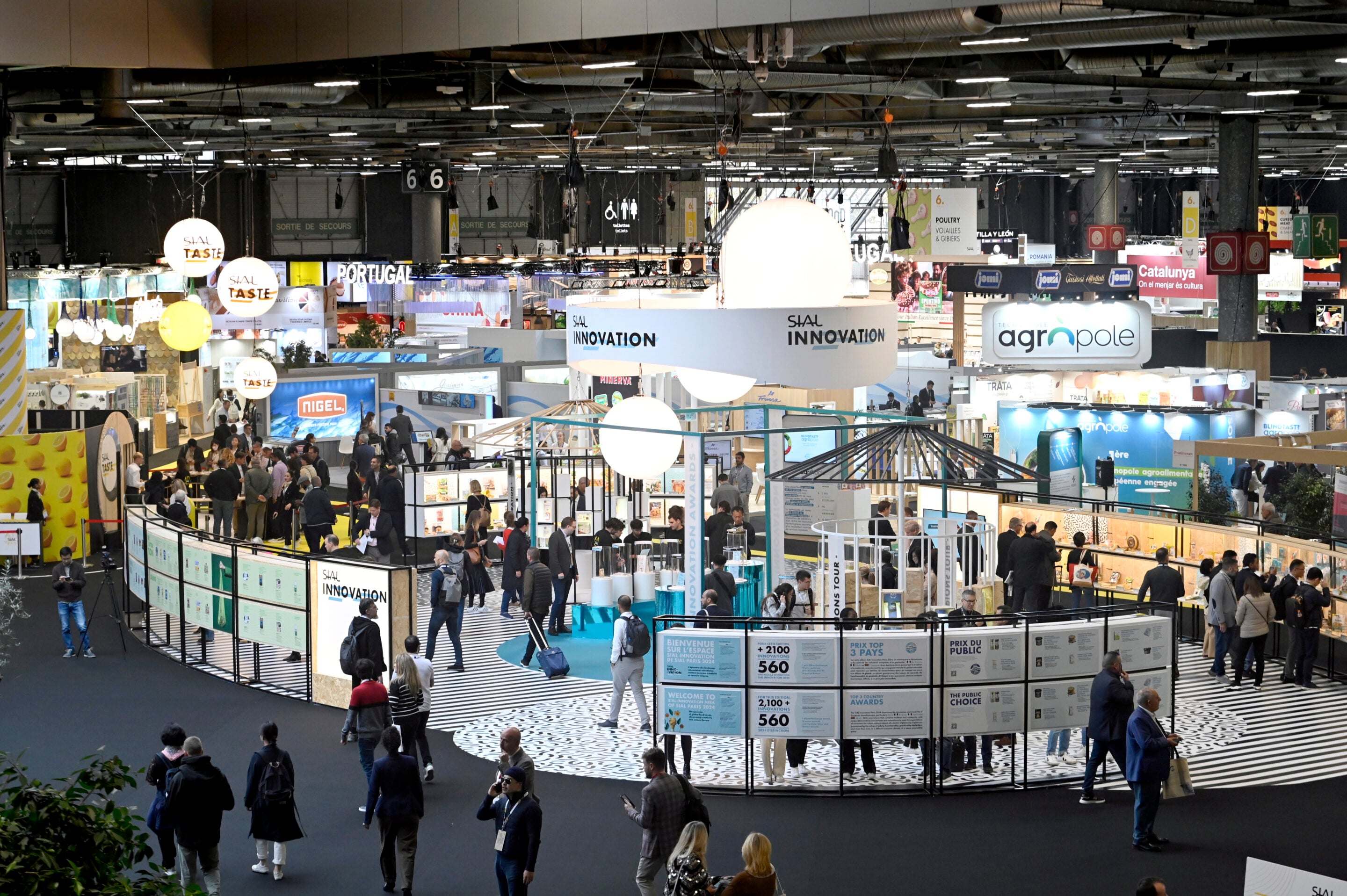 Large indoor trade fair with multiple stands, informational displays, and people walking and interacting in a well-lit exhibition hall. Signs with company names and bright overhead lights are visible.