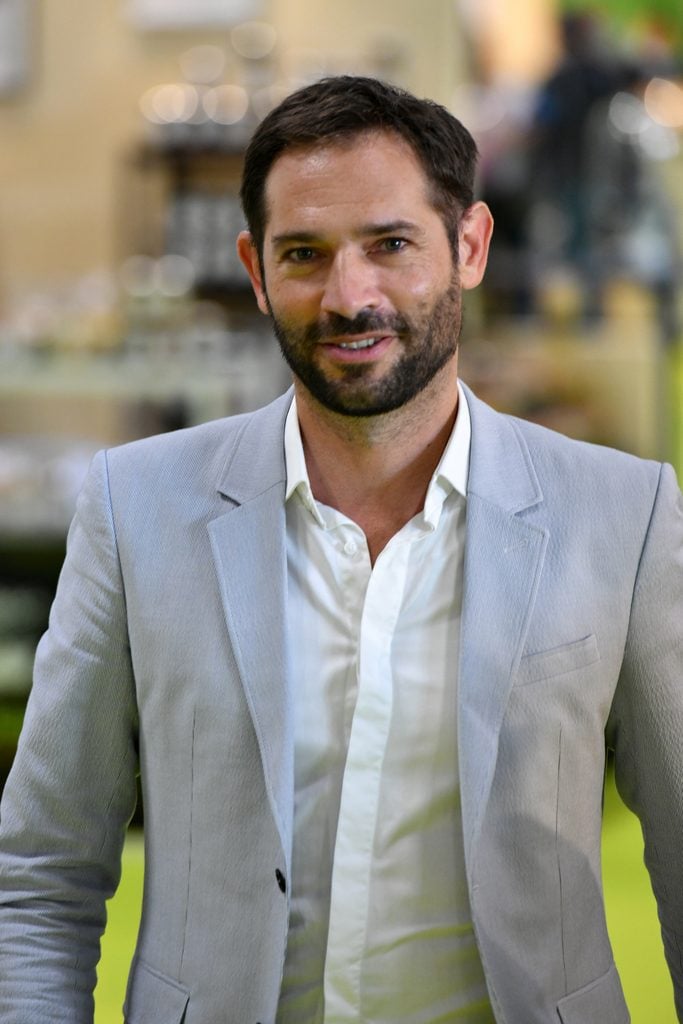A man with short dark hair and a beard wearing a light grey suit jacket and white shirt stands indoors, smiling at the camera.