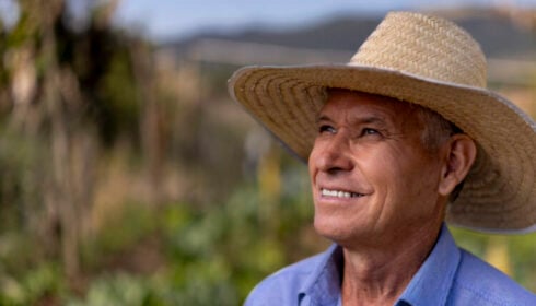 A smiling man in a straw hat and blue button-down shirt stands in a field.