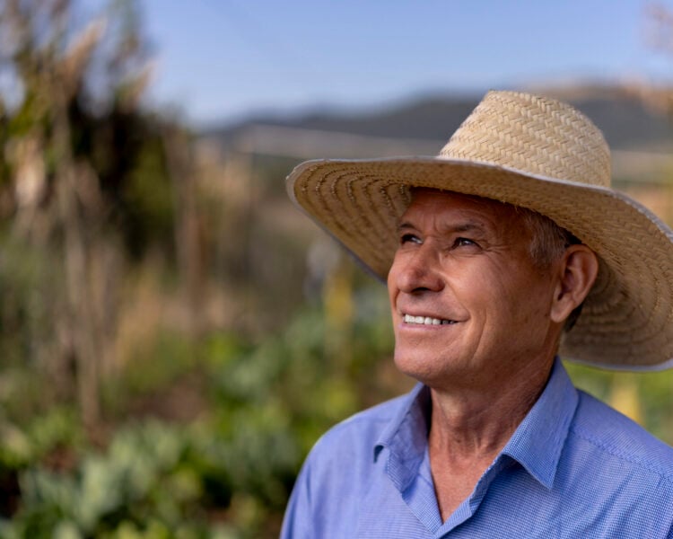 A smiling man in a straw hat and blue button-down shirt stands in a field.